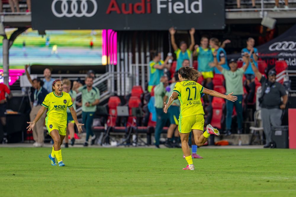 Soccer players in yellow uniforms celebrate a goal at Audi Field during a professional women's soccer match.