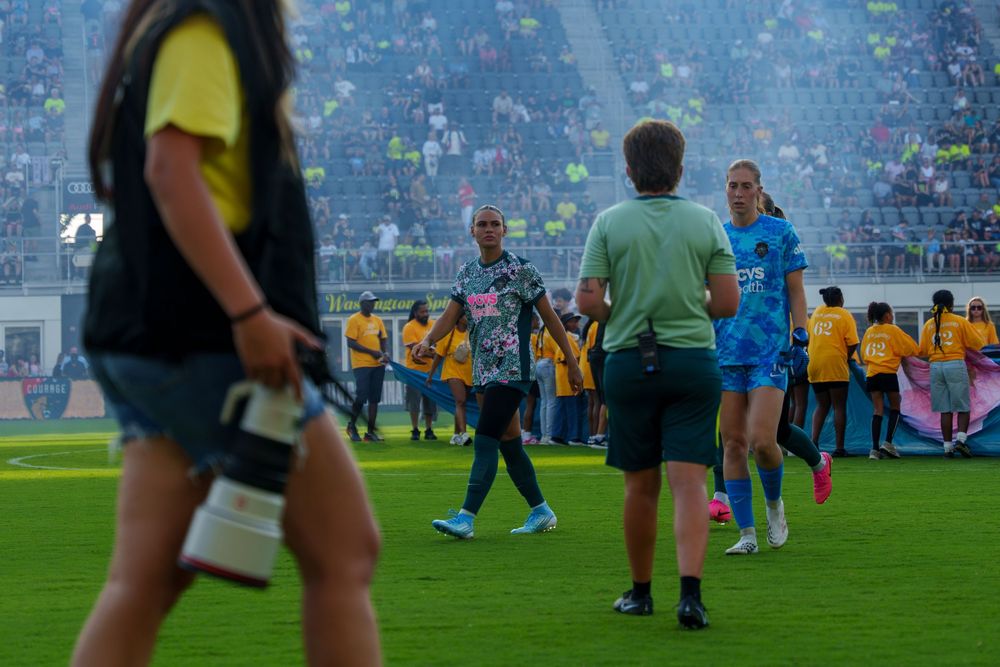 Soccer players and staff walk on a green field after a match, with fans visible in the stadium background.