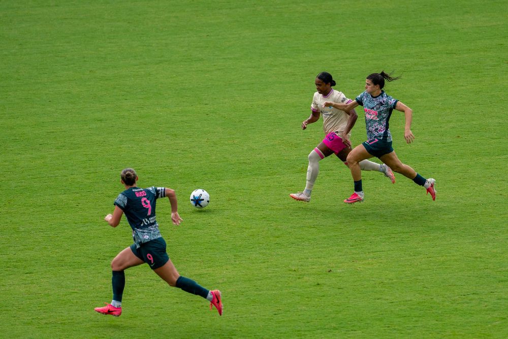 Female soccer players compete on a green field, with one player in beige dribbling past defenders in dark uniforms.