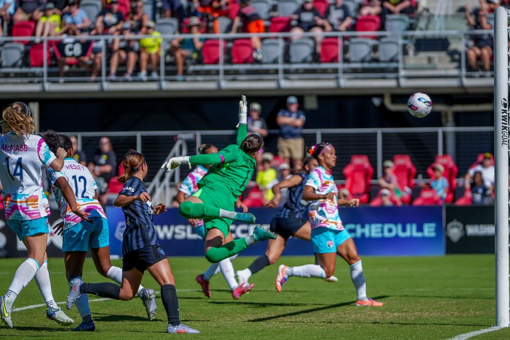 Soccer player in green goalkeeper uniform makes a diving save during a professional match with fans in the background.