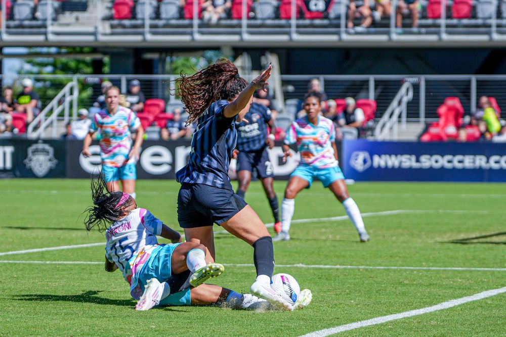 Soccer players compete for possession of the ball during an intense NWSL match on a sunny day.