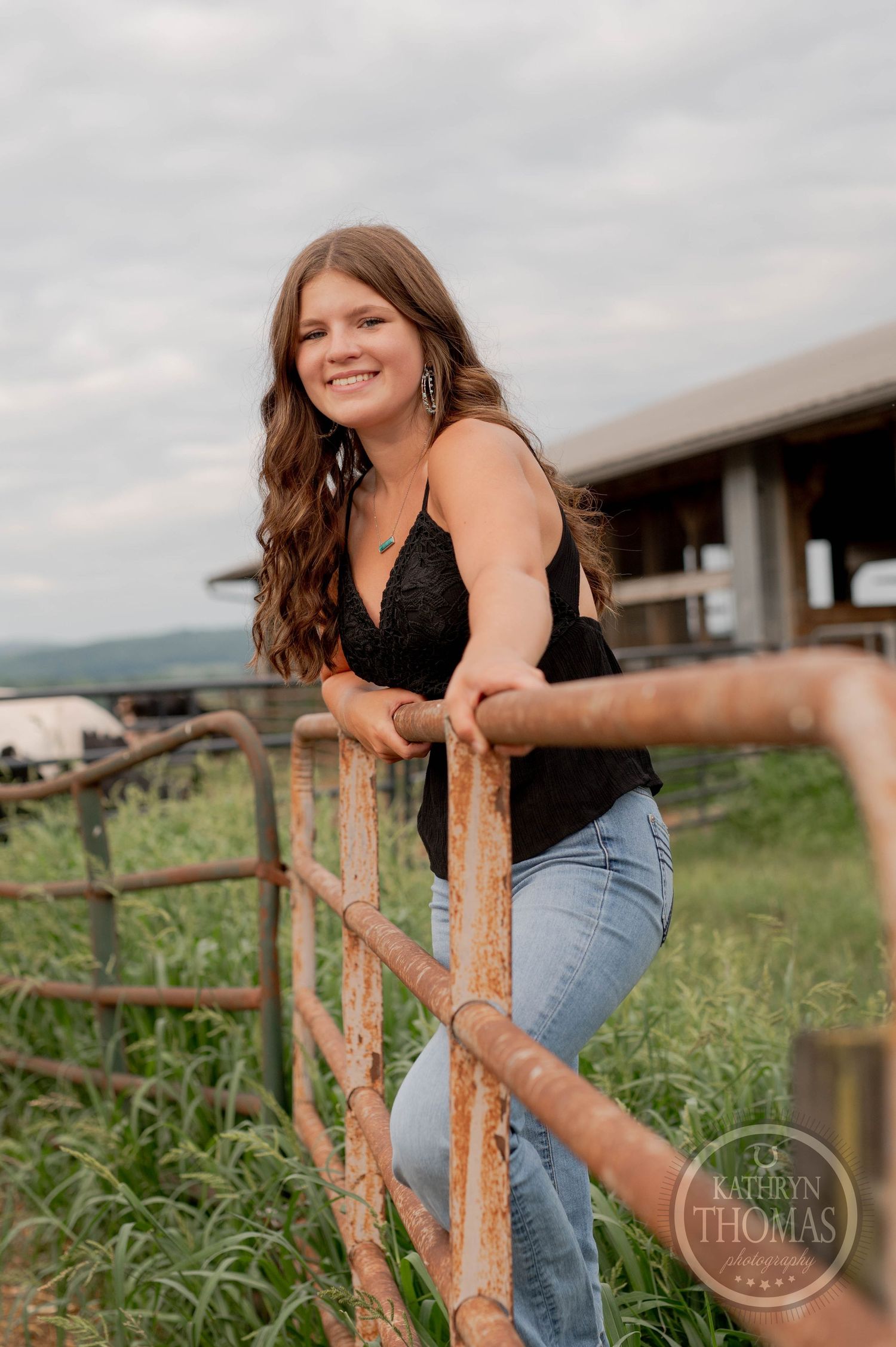 Woman in black tank top smiles while leaning on wooden farm fence under cloudy sky.