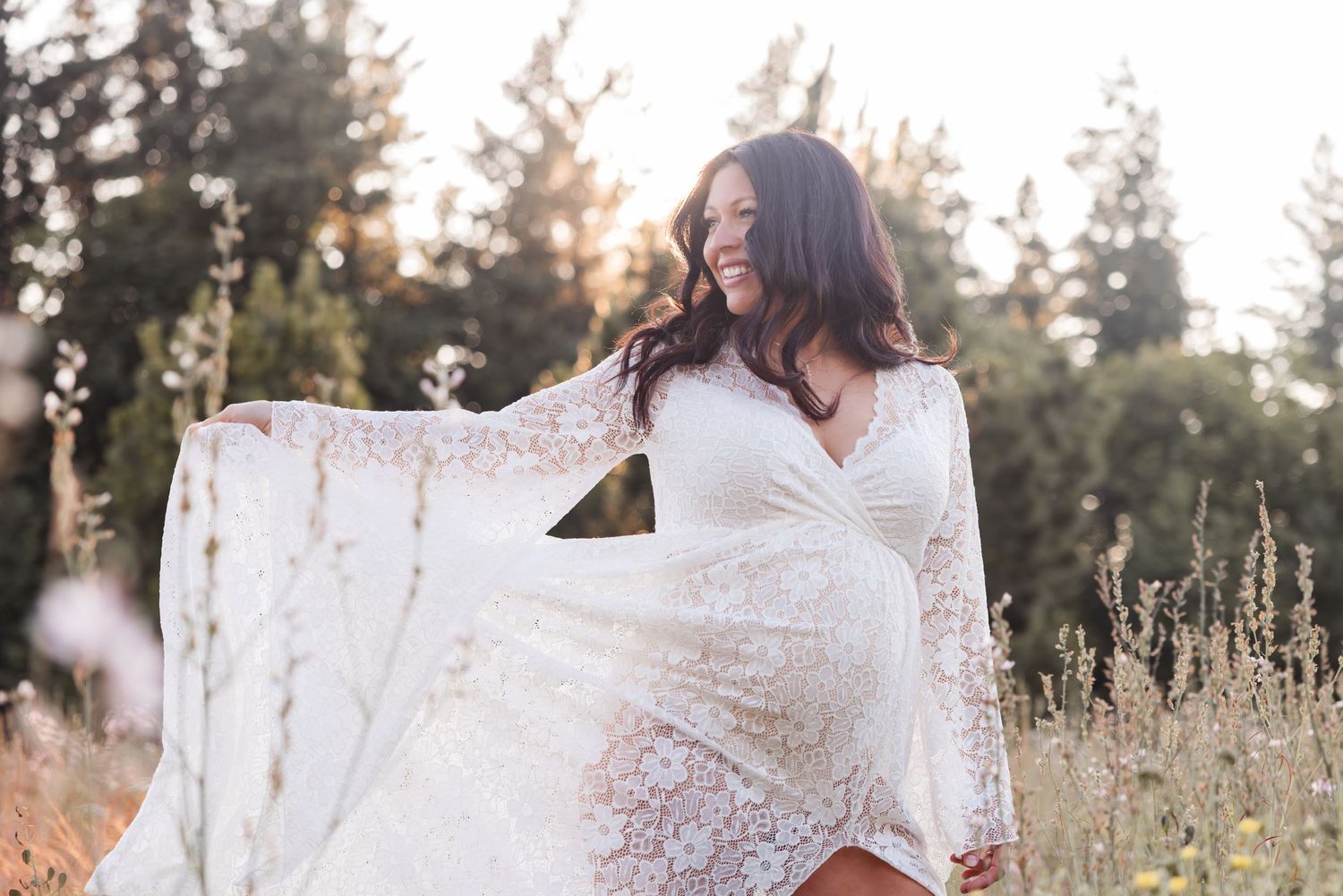 A white lace maternity dress flows in a golden field at sunset with soft sunlight filtering through trees.