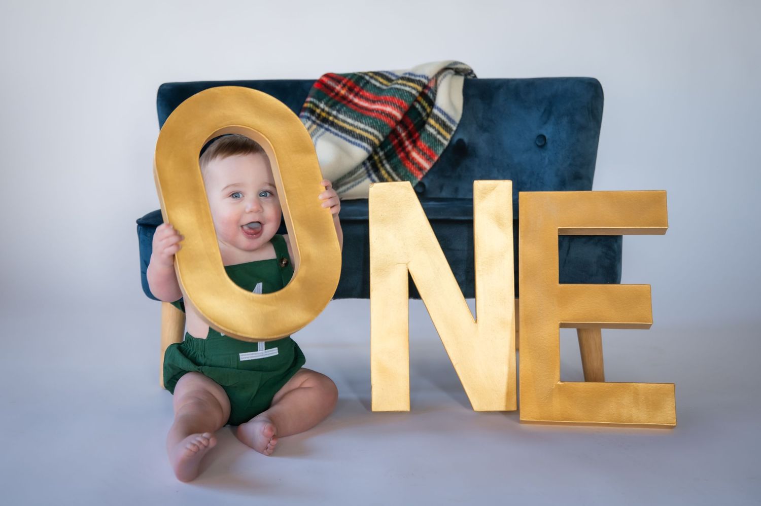 First birthday photo with large gold ONE letters against a blue tufted background in a studio setting.