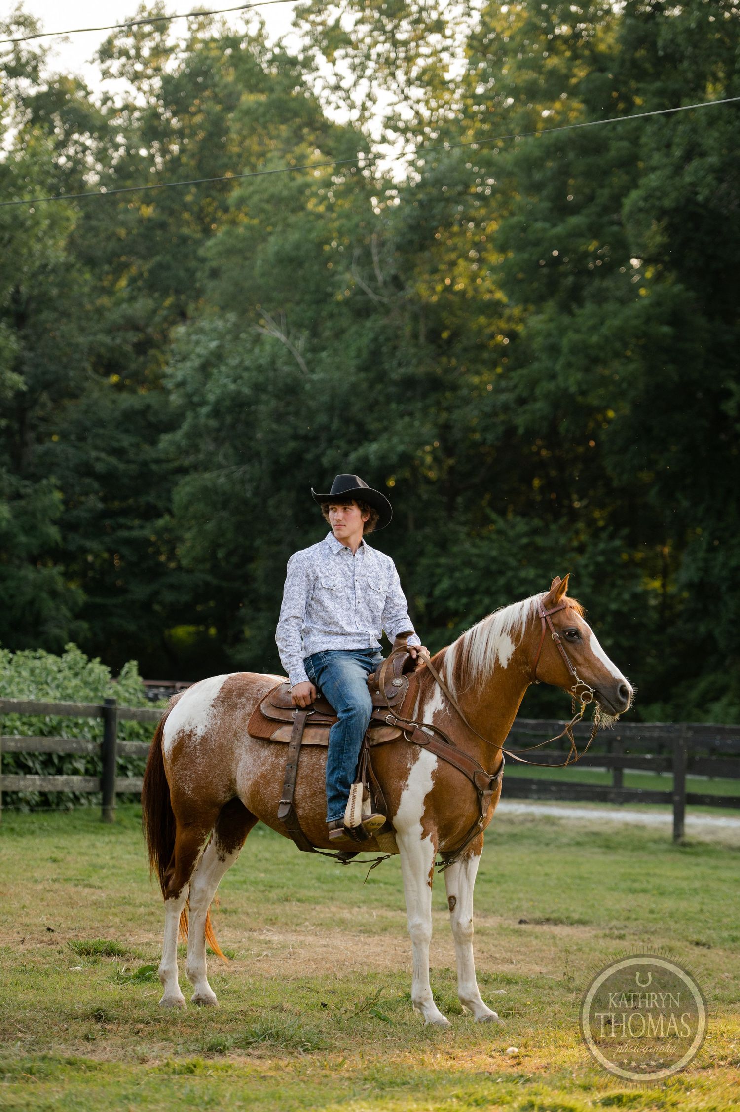 A paint horse with western saddle stands with rider in a fenced paddock.