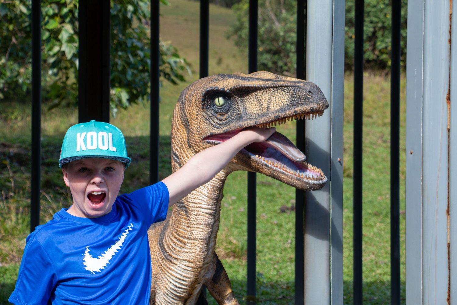 A person in a blue Nike shirt and teal cap poses excitedly next to a realistic velociraptor dinosaur model outdoors.