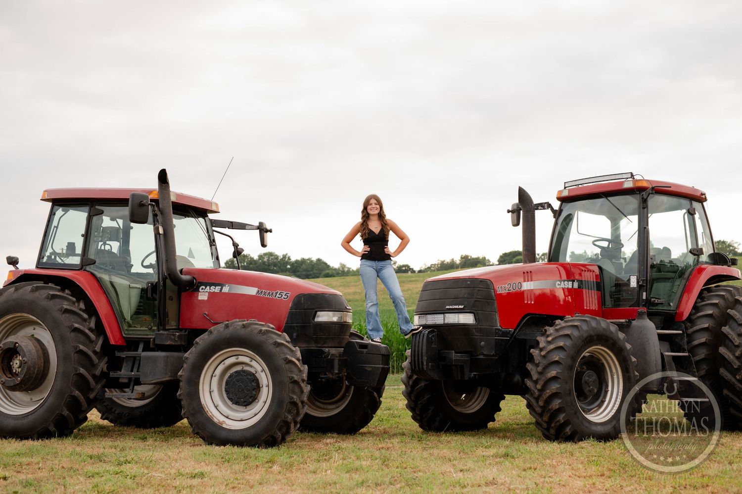Woman stands confidently between two large red farm tractors in rural field setting.