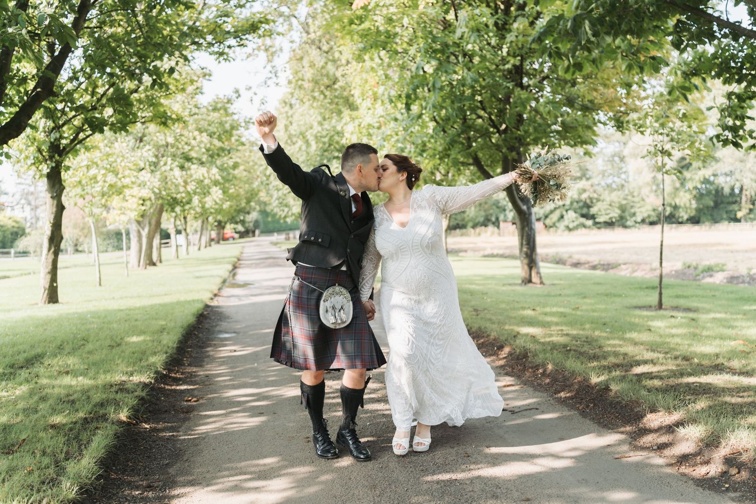 A newlywed couple walks joyfully down a tree-lined path, with the groom wearing a traditional Scottish kilt.