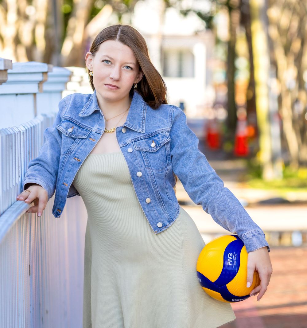 A person in a denim jacket holds a yellow and blue volleyball while leaning against a white fence outdoors.