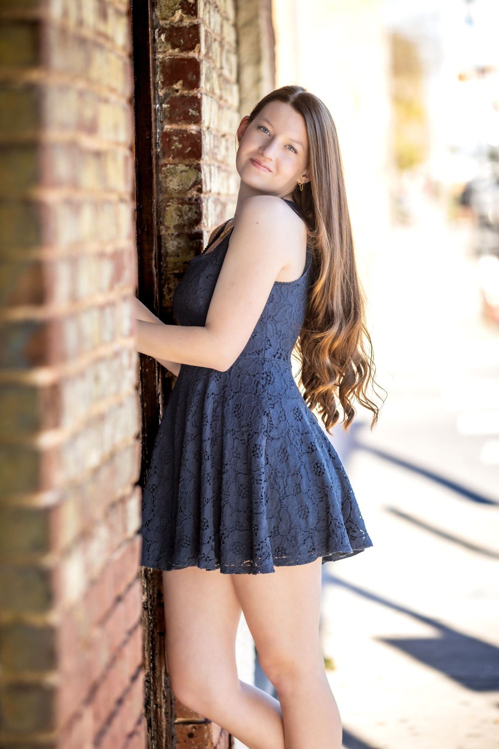A person in a navy blue lace dress poses beside a brick wall on a sunny day with soft lighting.