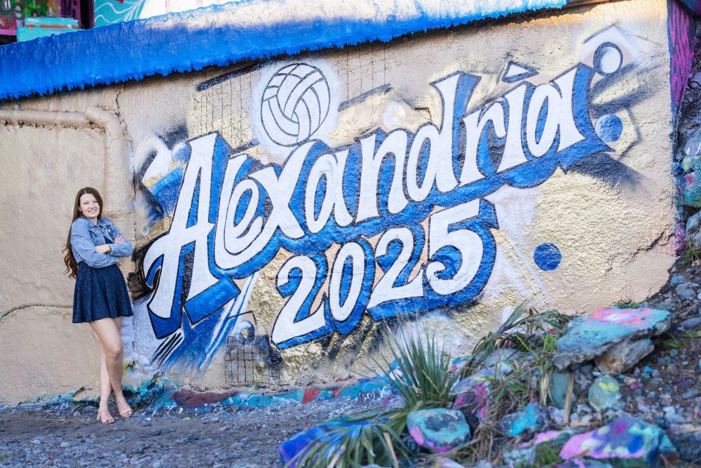 Blue and white graffiti mural reading 'Alexandrine 2025' painted on concrete wall with floral accents.