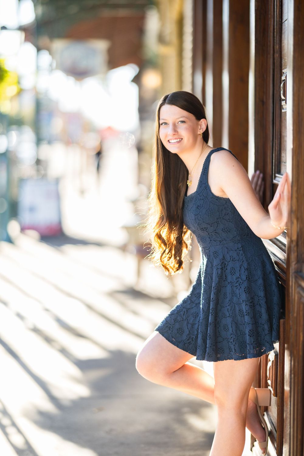 A woman in a navy blue lace dress poses on a sunny sidewalk against a wooden doorframe.