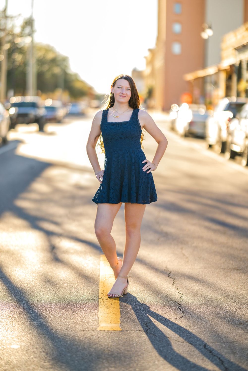 A person in a navy blue sundress poses on a sunlit street during golden hour while cars are parked along the curb.