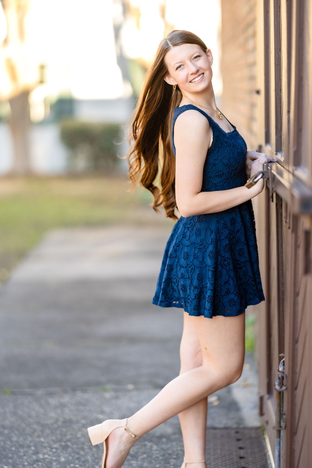 A graceful pose in a navy blue lace dress and nude heels next to a wooden wall during golden hour.