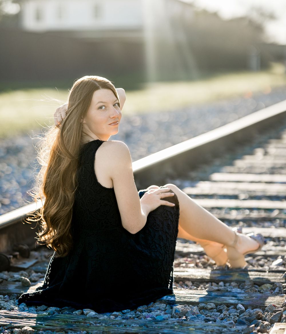 Figure in a black dress sits gracefully on train tracks during golden hour as sunlight creates a dreamy backlit effect.