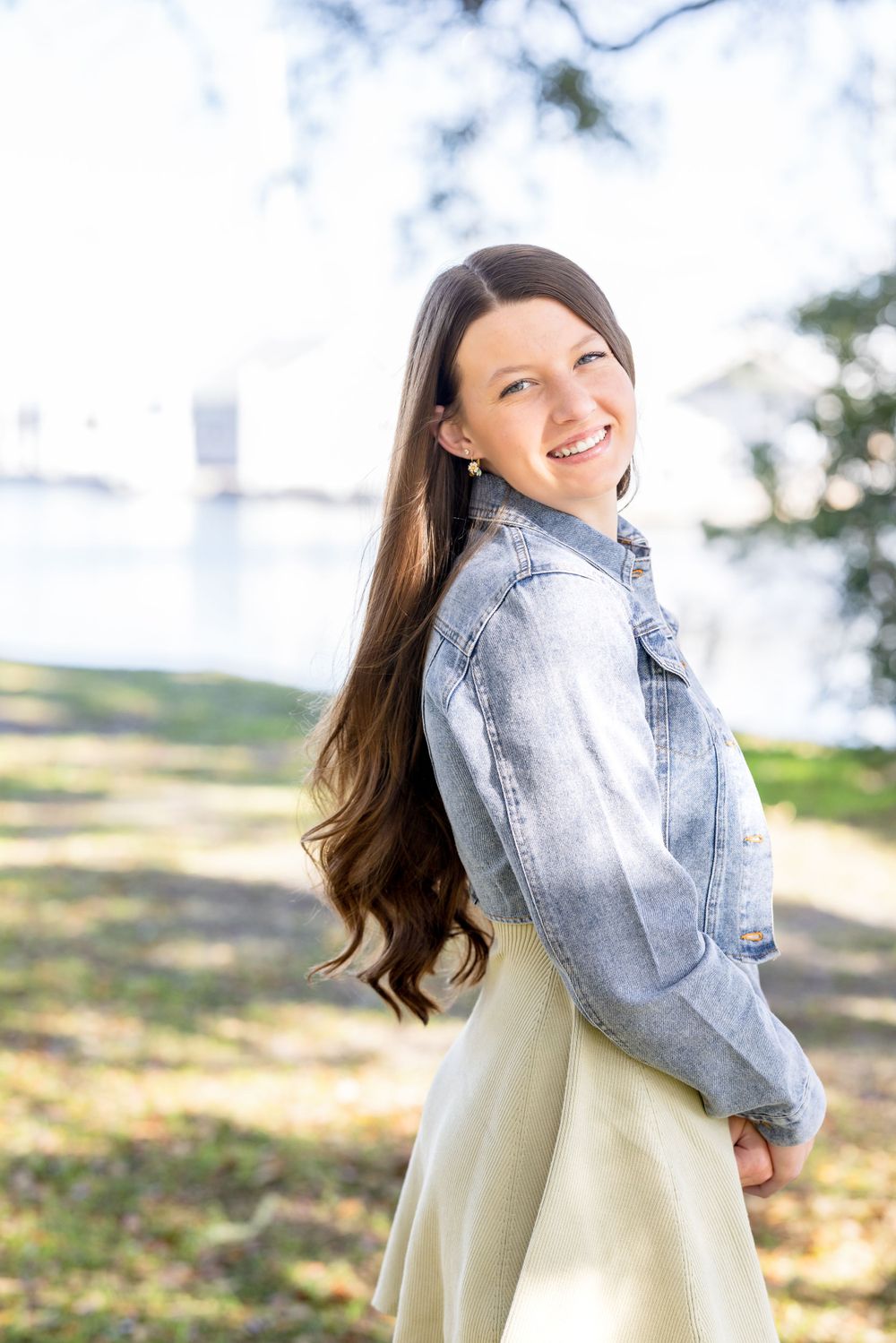 A woman in a light denim jacket and cream dress poses outdoors with a bright smile on a sunny day.