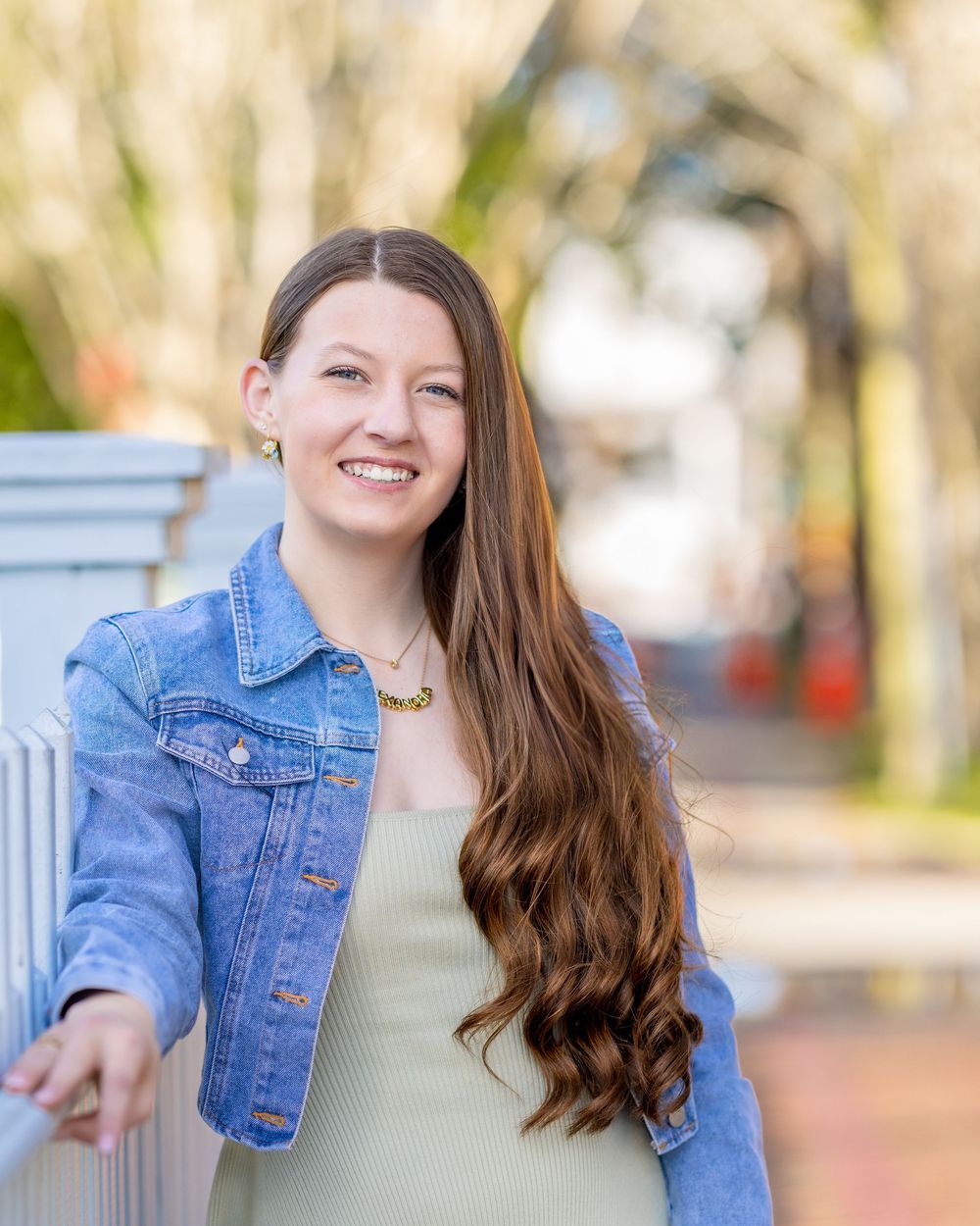 Smiling woman in denim jacket and beige top wearing long wavy brown hair stands outdoors by brick wall in natural lighting.