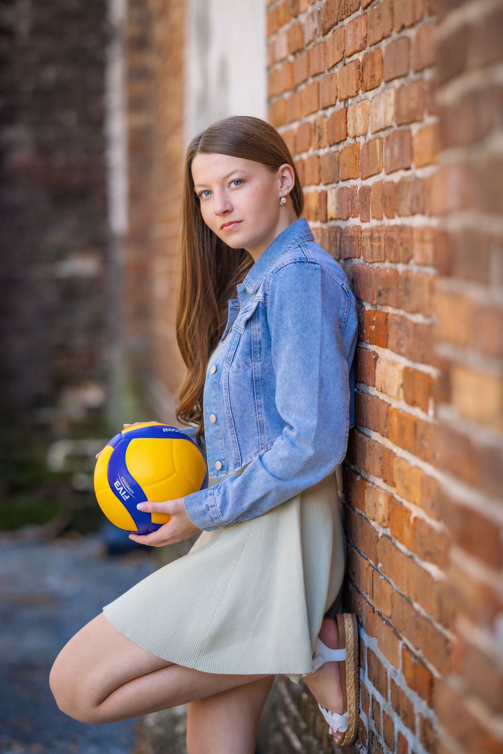 A person in a denim jacket and white dress leans against a brick wall while holding a blue and yellow volleyball.