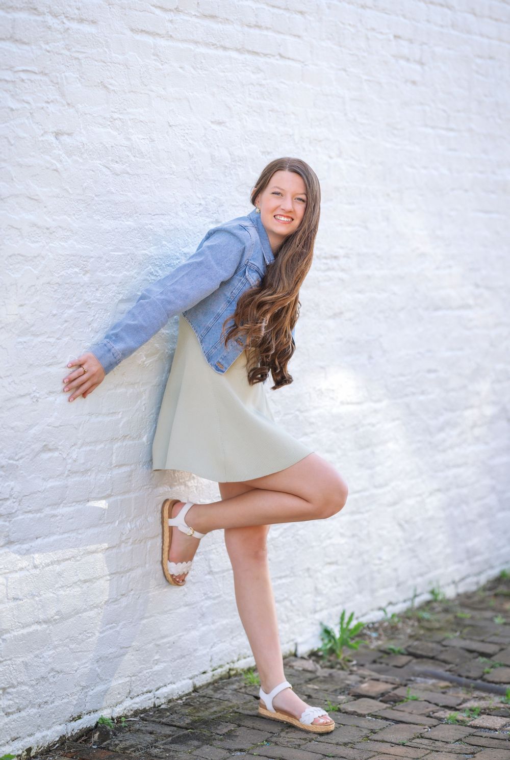 Joyful figure in denim jacket and flowy beige dress strikes playful pose against white brick wall outdoors.