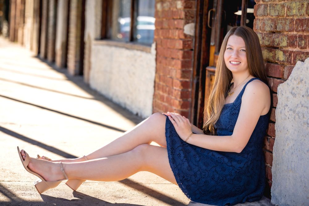 Someone wearing a navy blue dress sits against a brick wall in sunlight while smiling warmly at the camera.