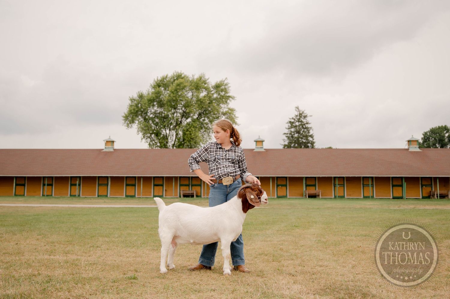 White goat and handler pose together in field with brick buildings in background.