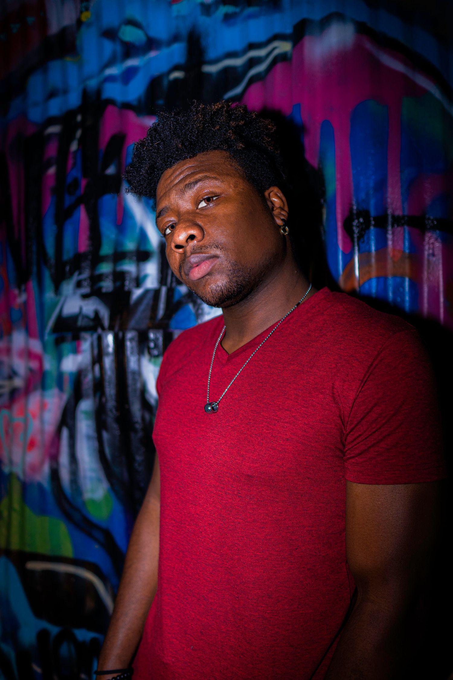 Portrait against colorful graffiti wall showing person in red t-shirt and necklace with dramatic lighting.