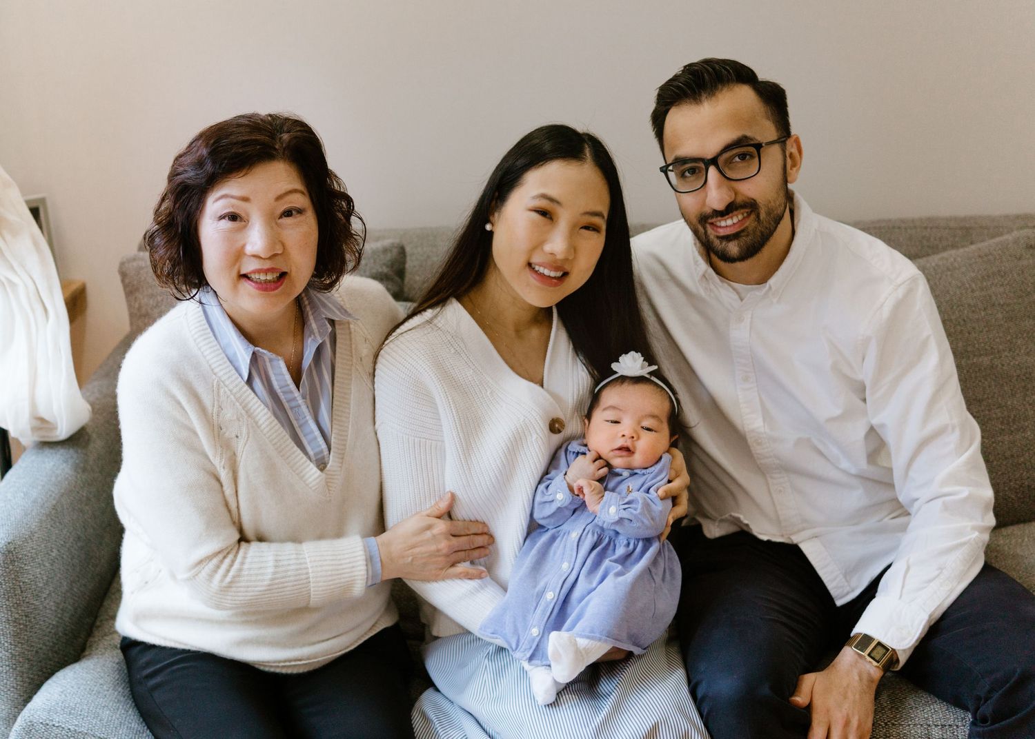 A family smiles together on a gray couch while holding a small baby wearing blue.