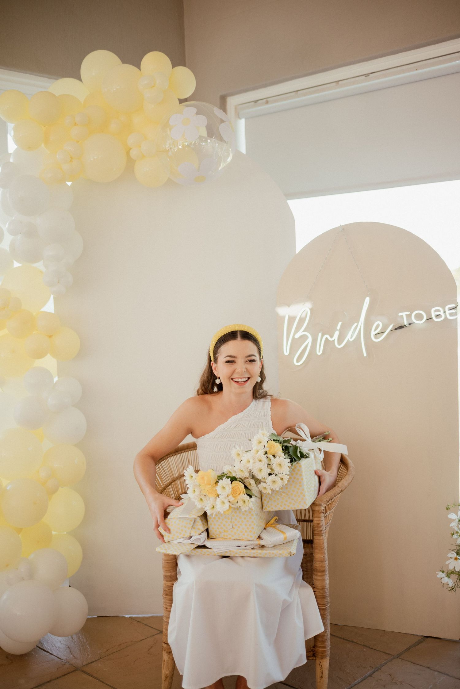 Bridal shower celebration with elegant white decor, yellow balloon arch, and 'Bride to Be' sign in a bright sunlit room.