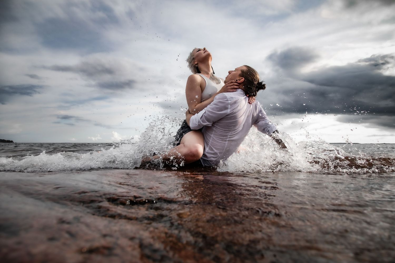 Romantic couple embracing in ocean waves under dramatic cloudy sky.