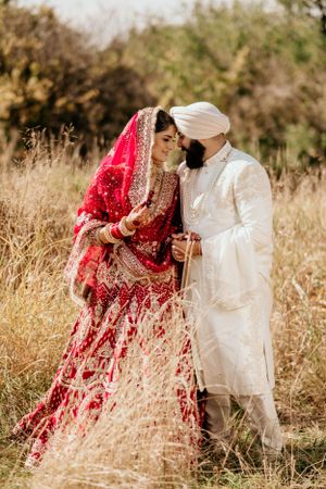Traditional South Asian wedding couple in red and white attire embrace in a sun-lit field of golden grass.