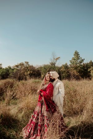 A couple in traditional Indian wedding attire poses together in a natural field setting during golden hour.