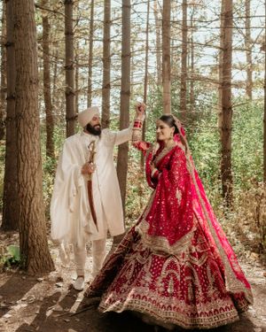 A couple in traditional Indian wedding outfits pose playfully among tall pine trees in a forest setting.