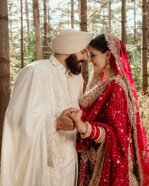 A couple in traditional wedding attire, with the bride in red embellished dress and groom in white, share an intimate moment in woods.