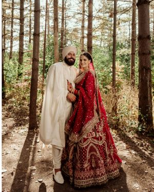 A couple wearing traditional red and white Indian wedding outfits poses together in a pine forest setting.