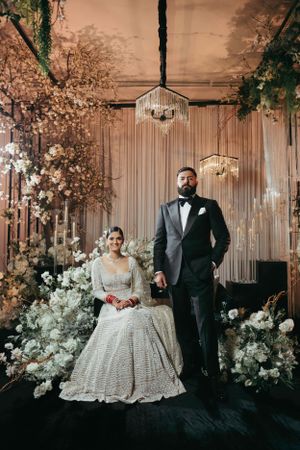 Elegant wedding couple stands against a luxurious backdrop of floral arrangements and crystal chandeliers in an indoor venue.