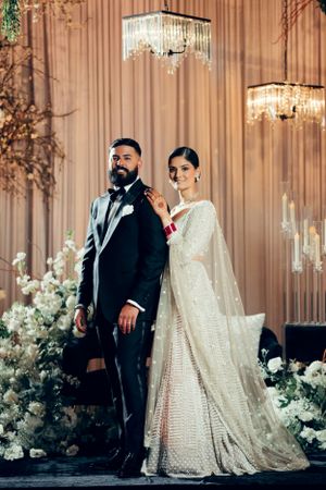 Elegant couple at wedding reception pose by floral decor and crystal chandeliers in traditional South Asian attire.