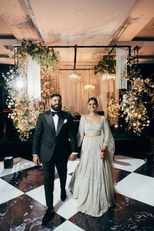 A couple stands together at their wedding ceremony with elegant floral decor and a checkered marble floor.