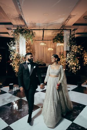 A newlywed couple walking hand in hand at their wedding reception decorated with floral arrangements and chandeliers.