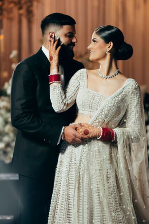 South Asian couple in traditional wedding attire sharing an intimate moment as bride wears white lehenga and groom wears black suit.