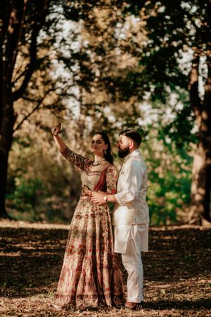 A couple in traditional Indian and Western fusion attire pose together outdoors in a sun-dappled forest setting.