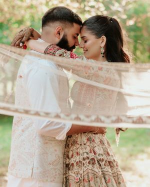 A romantic couple draped in sheer fabric share an intimate moment wearing traditional South Asian attire in an outdoor setting.