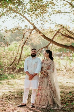 A couple in traditional South Asian wedding attire poses together under blooming trees in a romantic outdoor setting.
