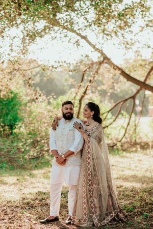 Couple in elegant Indian outfits pose together under blooming tree branches in natural outdoor setting.