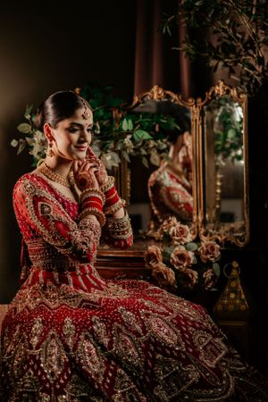 Beautiful bride in ornate red and gold lehenga poses elegantly with mirror reflection and floral decor in moody lighting.