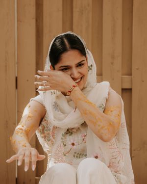 A joyful bride in a white outfit with henna adorned hands smiles and gestures against a wooden backdrop.