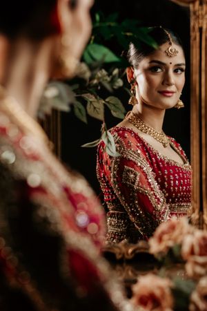 A reflection in the mirror shows an Indian bride wearing a red traditional lehenga with ornate gold jewelry and embellishments.