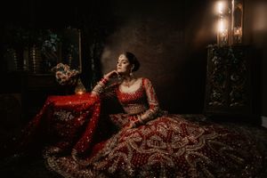 A bride in a red embroidered lehenga poses elegantly in dramatic moody lighting against a dark background.
