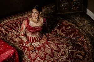 A bride in an ornate red and gold traditional Indian bridal lehenga sits gracefully on an intricately patterned carpet.