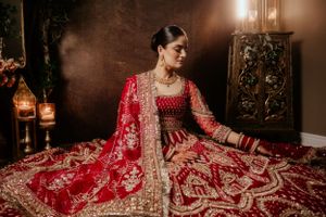 A traditional Indian bride wearing an ornate red and gold lehenga sits surrounded by glowing lanterns in a dimly lit room.