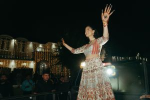A dancer in a white and red lehenga performs at an outdoor evening event with illuminated buildings in the background.